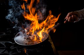 Side view mushroom frying with stove and fire and human hand in pan on black background