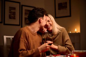 Happy young couple in love hugging, laughing, drinking wine, enjoying talking, having fun together celebrating Valentines day dining at home, having romantic dinner date with candles sitting at table.