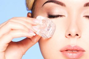 Closeup portrait of beautiful young woman applies the ice to face near eyes on a blue background.