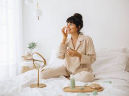 Happy woman doing routine skin care at home with beauty products. Woman sitting on bed at home and applying face cream.