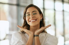 Cheerful business woman with glasses posing with her hands under her face showing her smile in an office. Playful hispanic female entrepreneur looking happy and excited at workplace