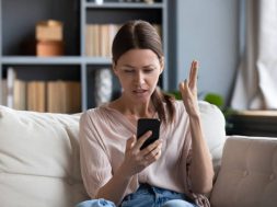 Confused angry woman having problem with phone, sitting on couch at home, unhappy young female looking at screen, dissatisfied by discharged or broken smartphone, reading bad news in message